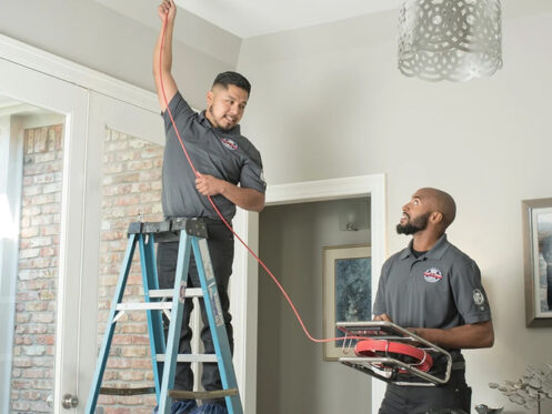Two men are cleaning out an air duct. One man is on a ladder, while the other is help hold the cleaning device.